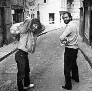 Two men stand on a narrow cobblestone street in a city. One has a bag slung over his shoulder and is looking back, while the other, holding items in his arms, smiles at the camera. The image is in black and white.